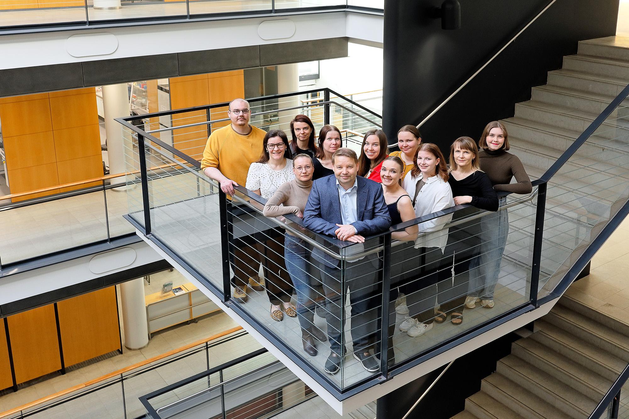 A group of thirteen people stands together on a stair landing in a modern indoor setting, surrounded by glass railings and natural light. They are smiling and appear relaxed, dressed in casual and business-casual attire.