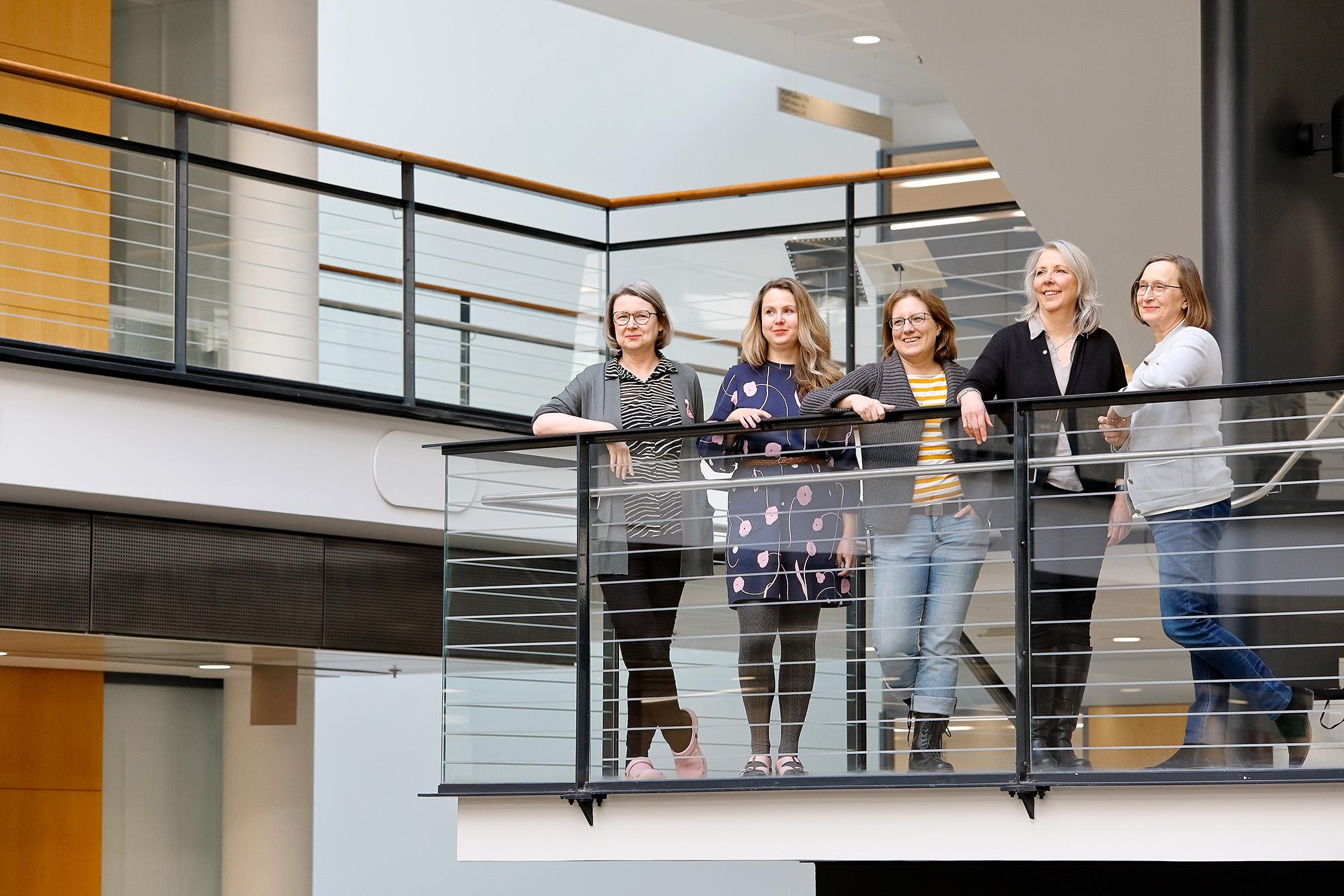 Five women stand together on an indoor balcony in a bright, modern building, smiling toward the camera. They are casually dressed and appear relaxed, suggesting a collegial and friendly work atmosphere.