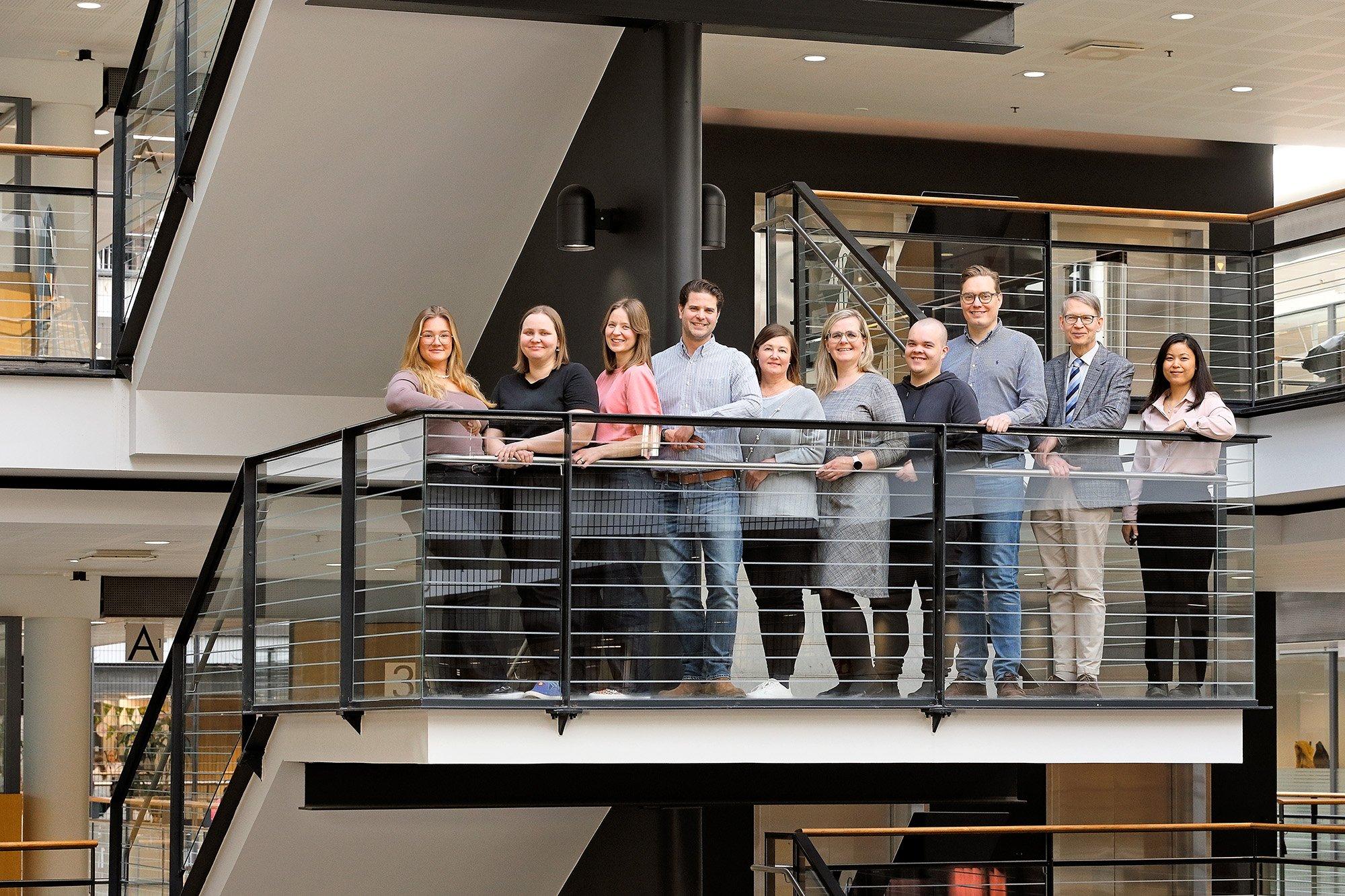 A group of twelve people stands on an indoor balcony in a modern, multi-story building, posing for a professional group photo. They are dressed in a mix of business and casual attire, smiling and standing close together in a well-lit setting.