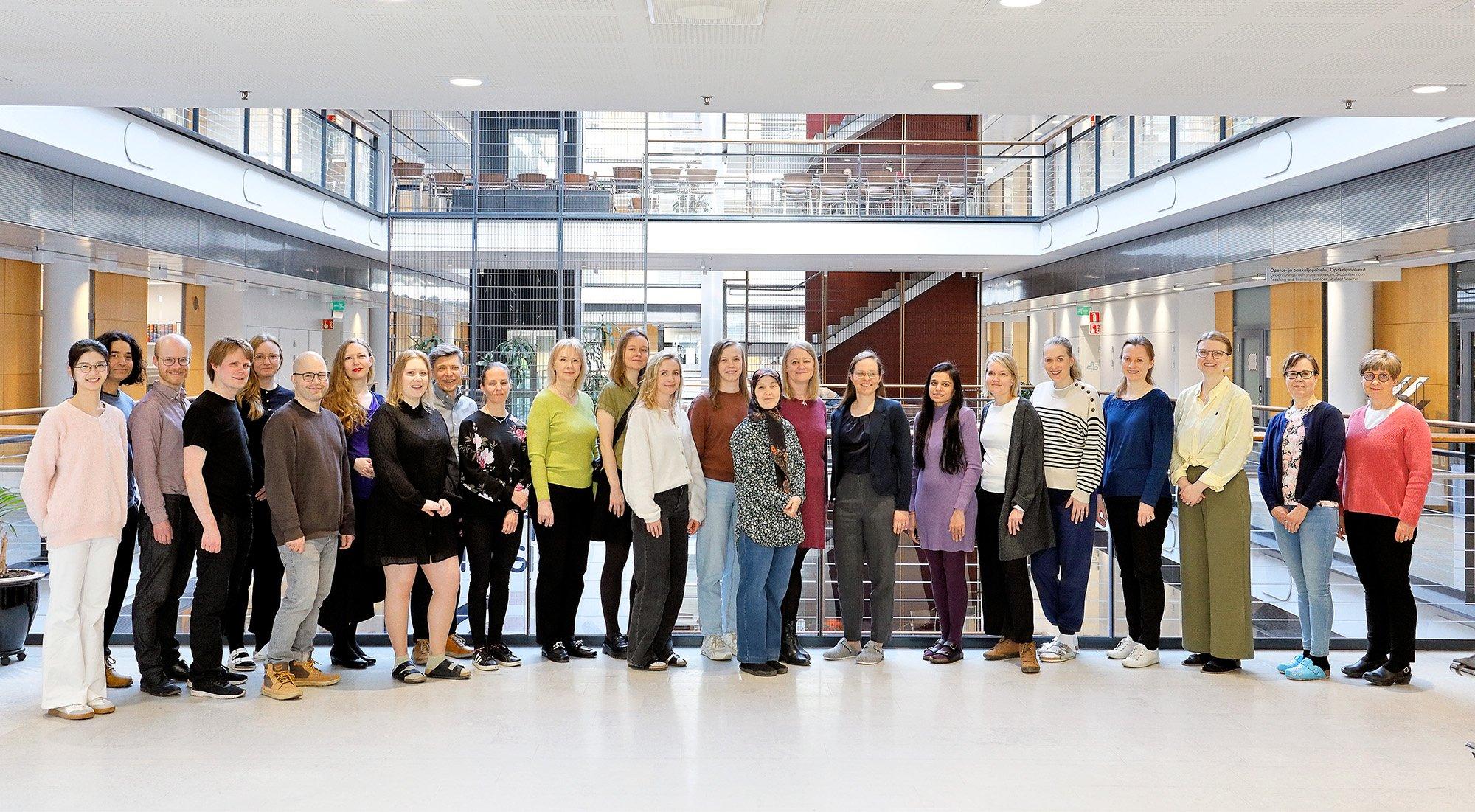 A large group of people stands in a bright, modern atrium, posing together for a group photo. The individuals are dressed in a variety of styles, smiling and facing the camera in a spacious, professional setting.