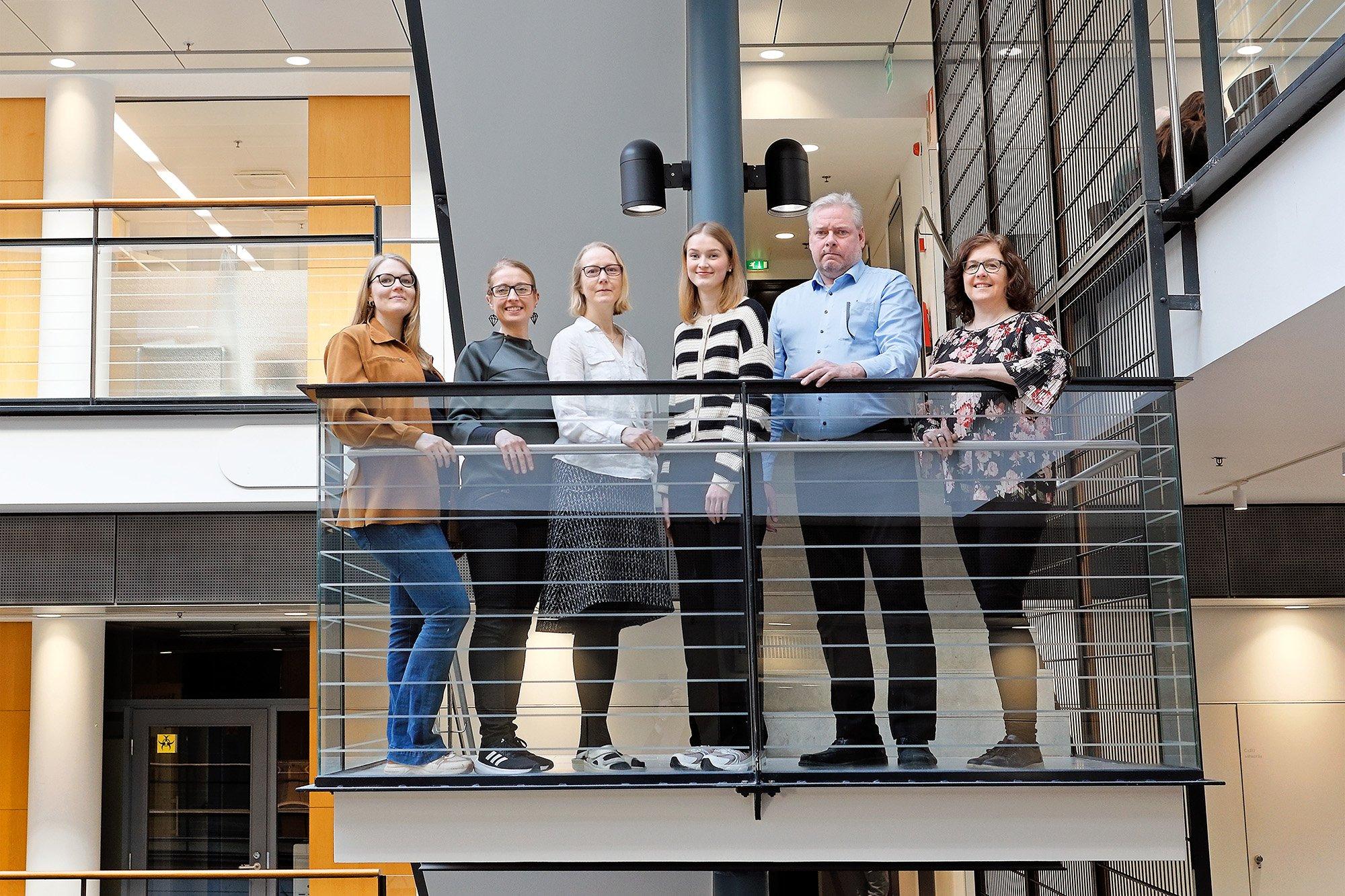 Seven people are standing on an indoor balcony in a modern office building, posing for a group photo in front of a staircase and glass railings. They are dressed in smart-casual attire and smiling, creating a warm and professional atmosphere.