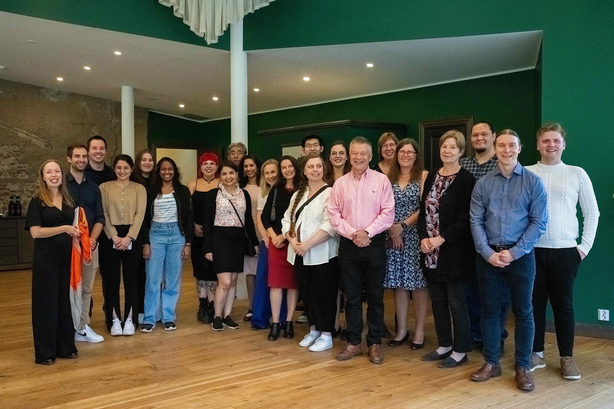 A diverse group of people is gathered in a warmly lit room with green walls, posing for a group photo. They are dressed in a mix of casual and formal attire, smiling and standing closely together.