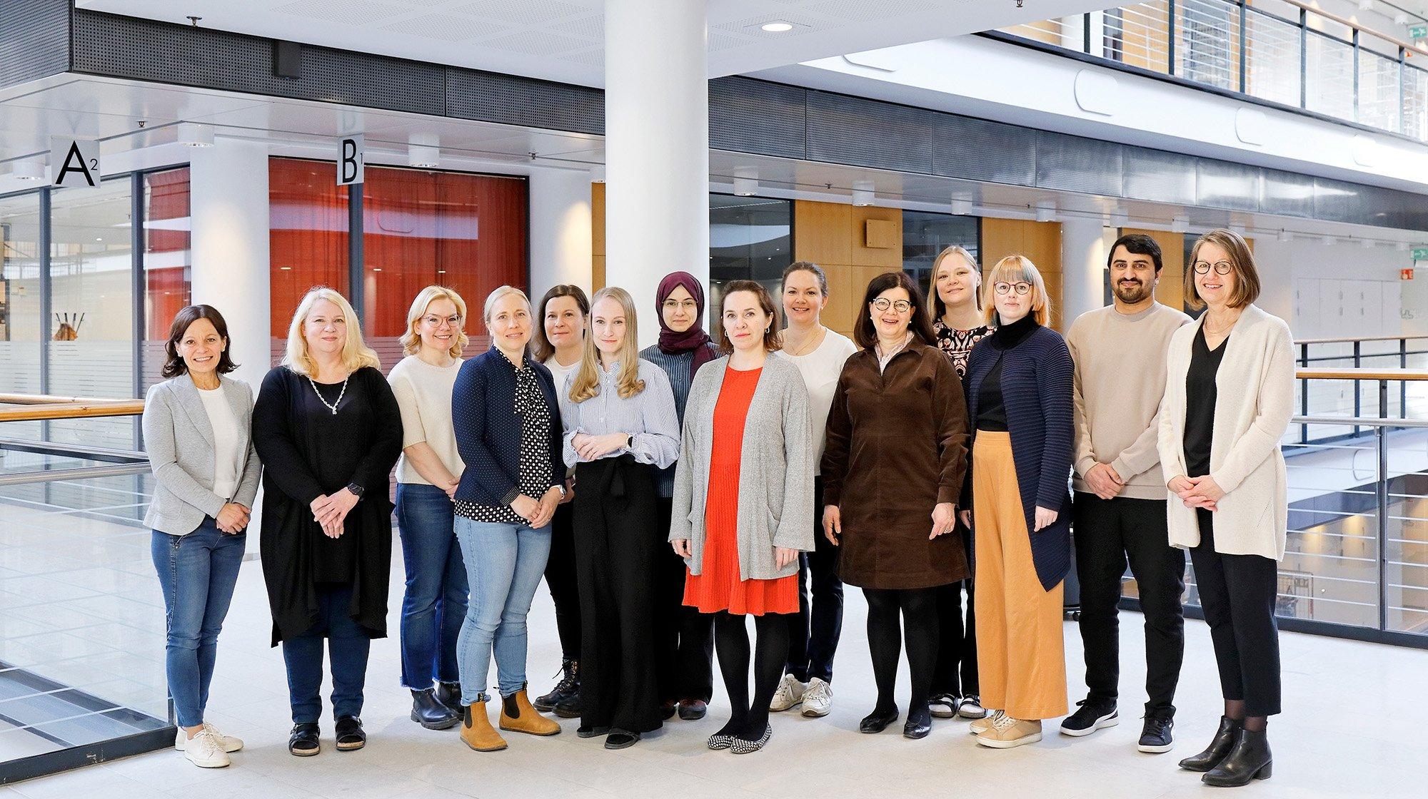 A diverse group of sixteen people stands in a bright, modern indoor space, posing for a group photo in front of red and wooden accent walls. They are dressed in casual and business-casual attire, smiling warmly in a professional yet relaxed atmosphere.