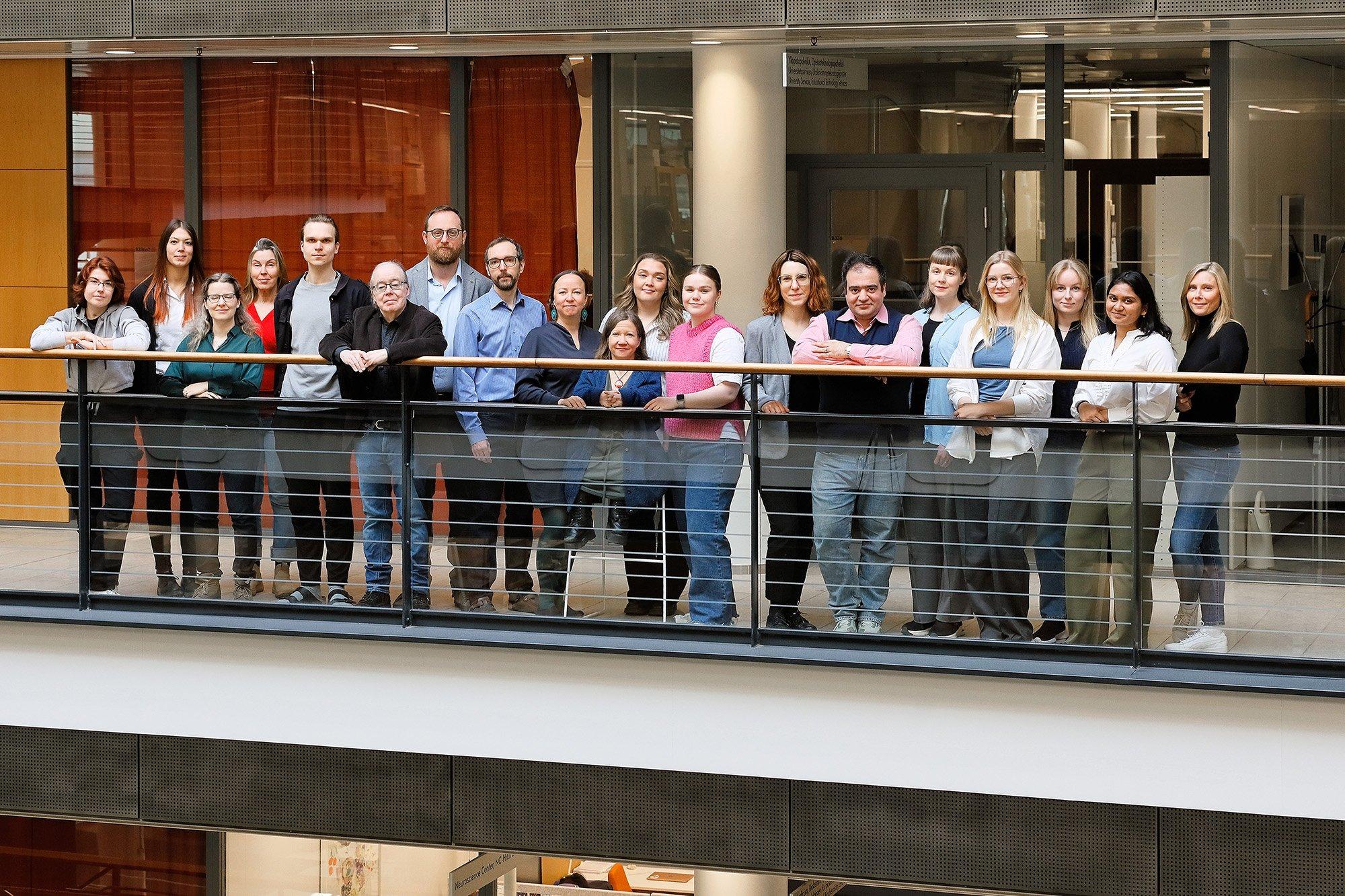 A group of around 20 people is standing side by side on an indoor balcony, posing for a team photo in a modern building with glass walls and warm wood accents. They are smiling and dressed in a mix of casual and professional attire.