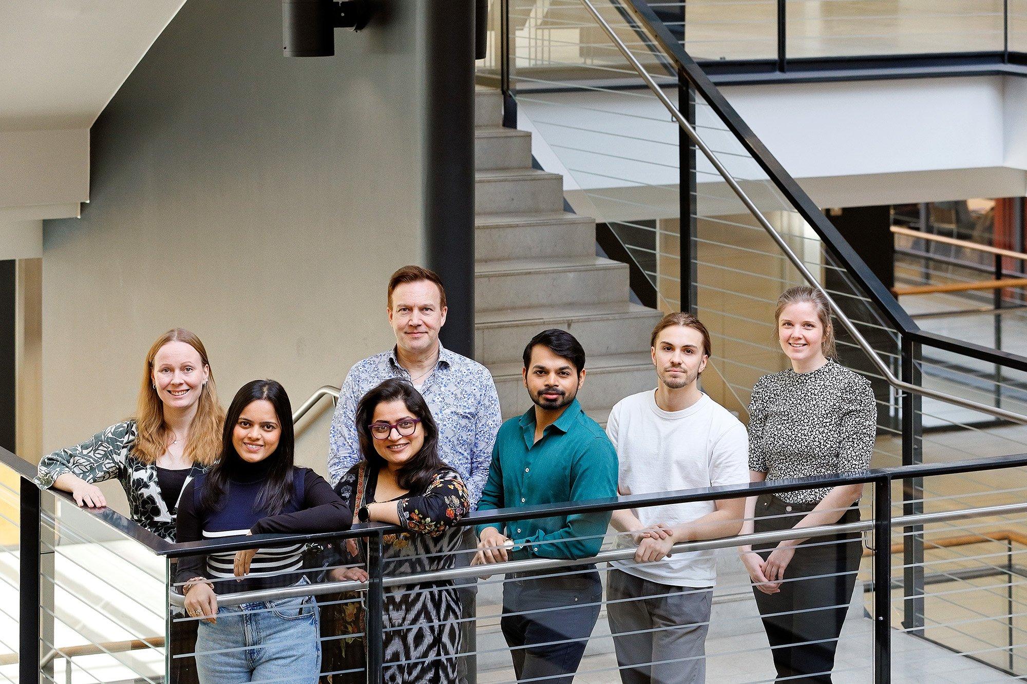 Seven people are standing together on a stair landing in a modern indoor space, posing for a group photo with a mix of relaxed and cheerful expressions. They are dressed in casual to smart-casual clothing, creating a warm and diverse team atmosphere.