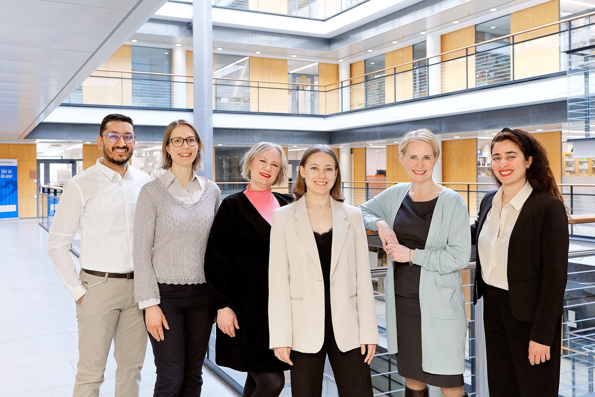 Seven people are standing side by side in a bright, modern indoor setting with glass railings and wood-accented walls. They are dressed in business and smart-casual attire, smiling confidently.