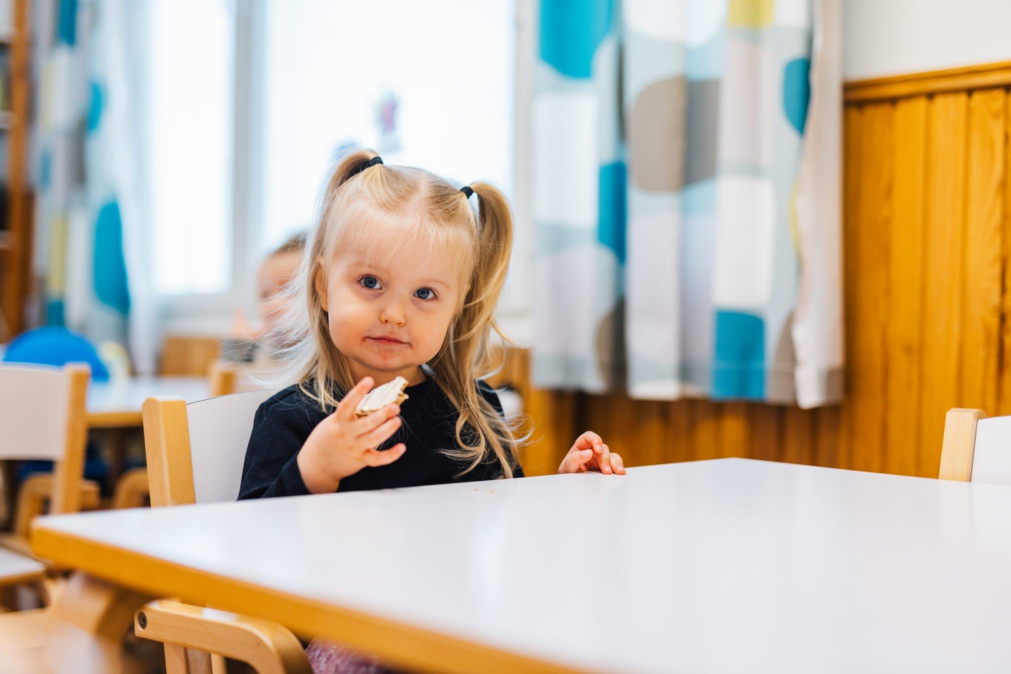 A young girl with blonde pigtails is sitting at a table in a daycare setting, enjoying a snack. She looks directly at the camera with a curious expression, while the warm and colorful environment surrounds her.