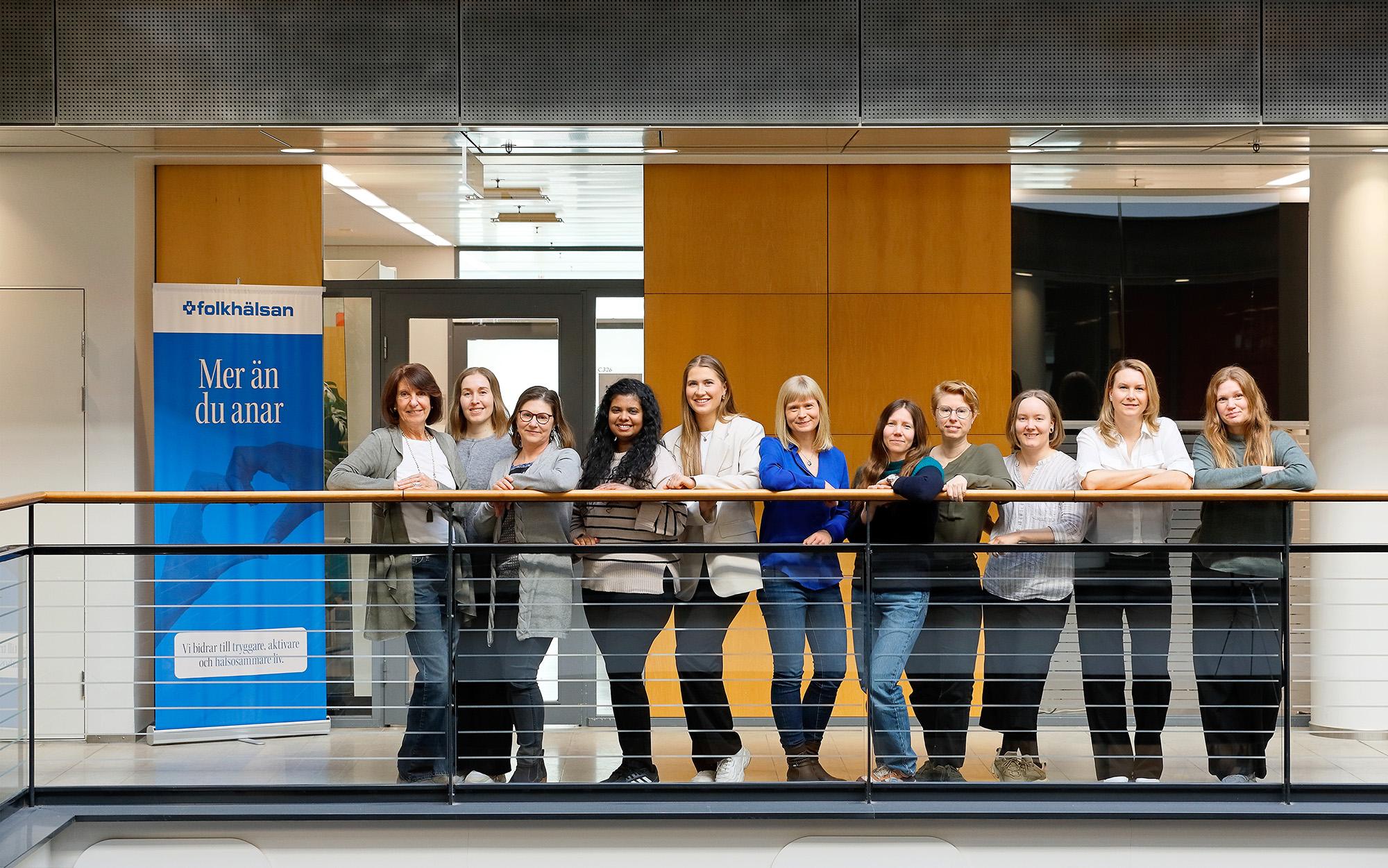 Twelve women are standing in a row on an indoor balcony in a modern building, smiling and posing confidently for a group photo. A Folkhälsan banner in the background adds a professional context to the friendly and collaborative atmosphere.