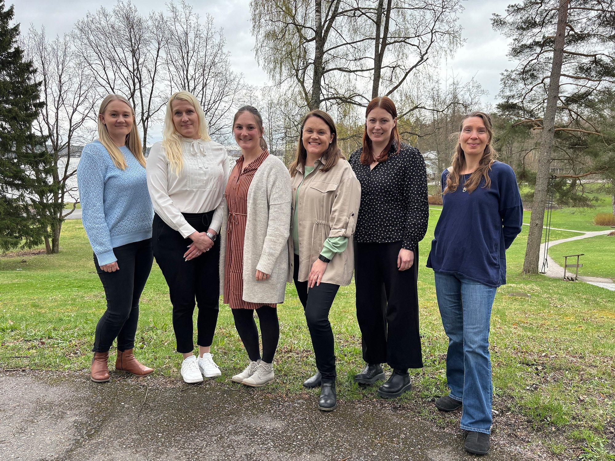 Six women are standing outdoors on a slightly overcast day, posing together and smiling in front of a scenic background with trees, grass, and a lake. They are dressed in casual to business-casual clothing, in a relaxed yet professional gathering.