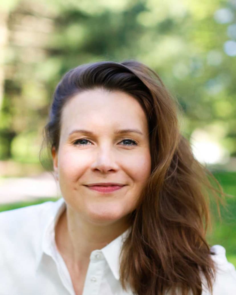 A woman with long brown hair and a white collared shirt is smiling softly at the camera in an outdoor setting. The background is blurred with green trees and sunlight, creating a warm and natural atmosphere.