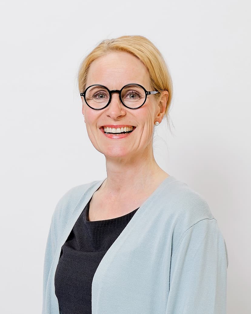 A smiling woman with blonde hair and round black glasses stands against a plain white background. She is wearing a light blue cardigan over a dark top, giving a professional and approachable appearance.