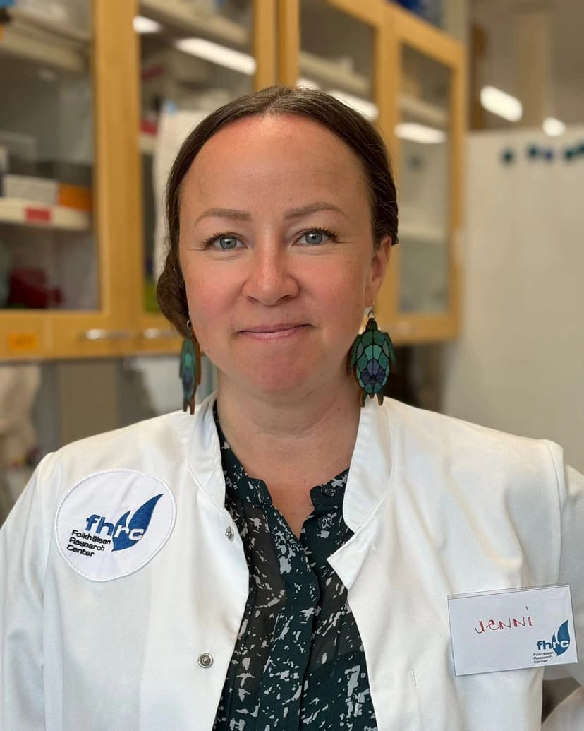 A woman wearing a white lab coat with a Folkhälsan Research Center logo stands in a laboratory, looking confidently at the camera. She has light eyes, wears patterned earrings, and a name tag that reads "Jenni".