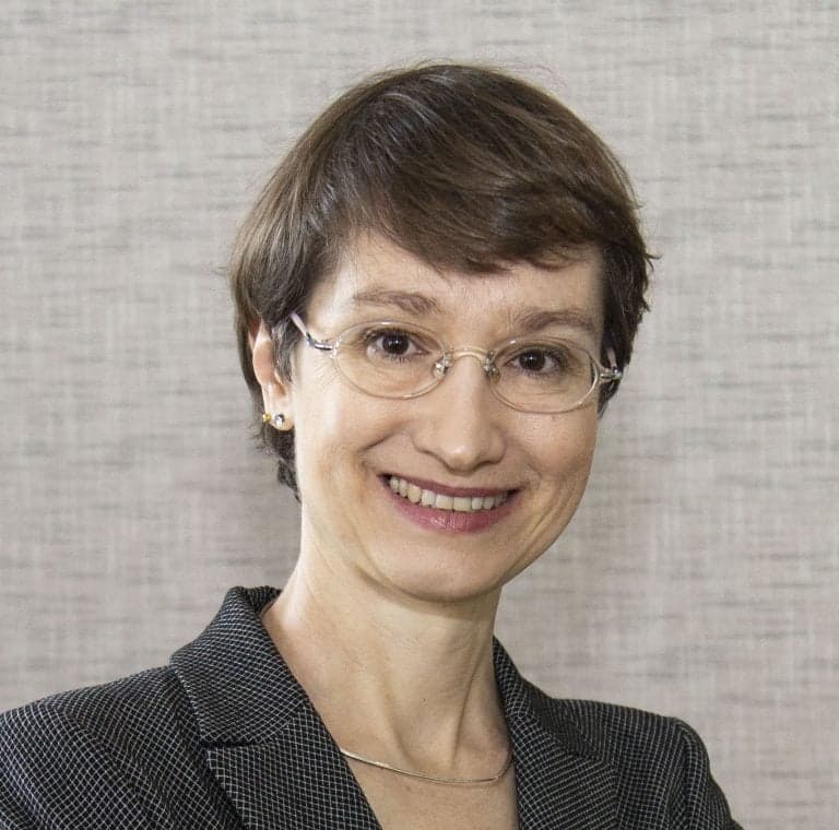 A woman with short brown hair, glasses, and a warm smile is dressed in a dark blazer with a subtle pattern. She is posing against a neutral textured background, looking directly at the camera.